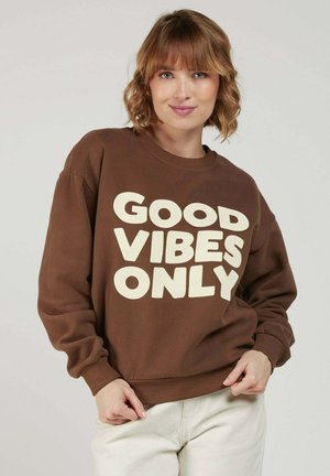 Jeune femme aux cheveux courts et ondulés porte un sweat-shirt marron avec un texte blanc en gras « GOOD VIBES ONLY », posant devant un fond uni.
