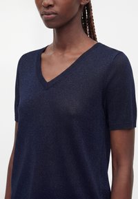 Femme portant un haut en maille bleu marine à col en V et manches courtes avec un motif texturé subtil, cheveux tressés visibles à l'arrière.