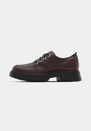 Chaussure en cuir bordeaux avec lacets noirs, bout rond, œillets dorés et finition texturée. Semelle en caoutchouc noir robuste avec des détails de traction.