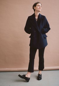 Manteau bleu marine croisé avec des poches latérales, porté sur un haut noir et un pantalon ajusté, associé à des chaussures à plateforme noires sur un sol gris.