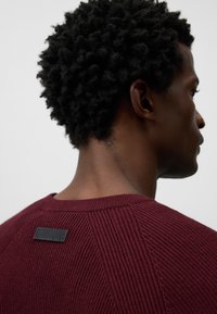 Burgundroter Strickpullover mit gerippter Textur, rundem Ausschnitt und einem schwarzen Etikett am Nacken.