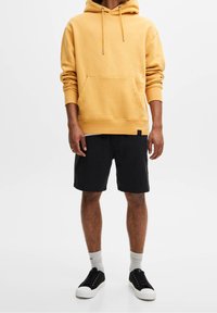 Persona de pie, vestida con una sudadera amarilla, pantalones cortos negros y zapatillas negras con suelas blancas. El fondo es blanco liso.