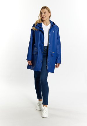 Blauwe waterdichte regenjas met een capuchon, voorzien van drukknoopsluitingen en twee voorzakken. Gekocht over een wit shirt en blauwe spijkerbroek.