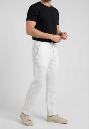 Uomo con indosso una maglietta nera a maniche corte, pantaloni bianchi con coulisse e sneakers beige, in piedi contro uno sfondo chiaro e uniforme.