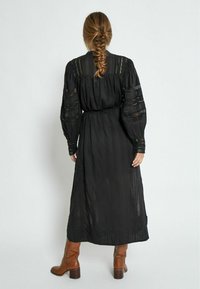 Black maxi dress with long, puffed sleeves and a fitted waist, featuring intricate lace details and a textured fabric. Paired with brown heeled boots.