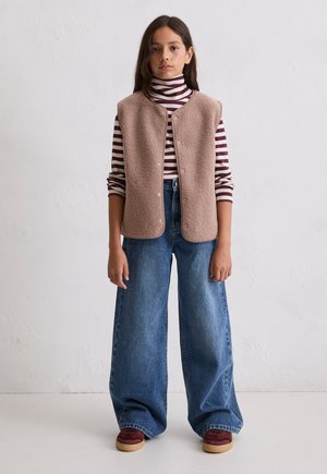 Meisje draagt een beige fleecevest met knopen over een bordeauxrood-wit gestreepte coltrui, wijde blauwe jeans en bordeauxrode sneakers.