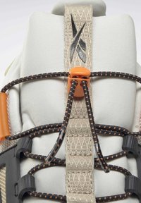 Primer plano de la lengua de una zapatilla con tela beige, cordones elásticos negros y naranjas, y un tope de cordón de plástico naranja en la parte superior.