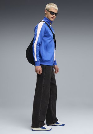 Blå jakke med hvide striber, båret med sorte, loose-fit jeans og hvide sneakers med blå detaljer. Modellen har kort, lyst hår.