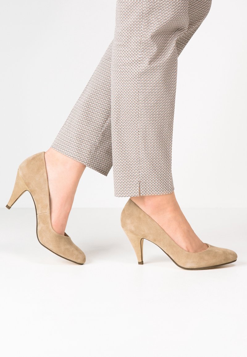 Pieds portant des chaussures à talons hauts en daim beige associées à un pantalon clair à motifs, longueur cheville, sur fond blanc.