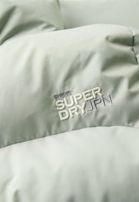 Polstret jakke i lysegrønt stof med en glat tekstur, der præsenterer det hvide broderede logo "SUPER DRY JPN" fremtrædende.
