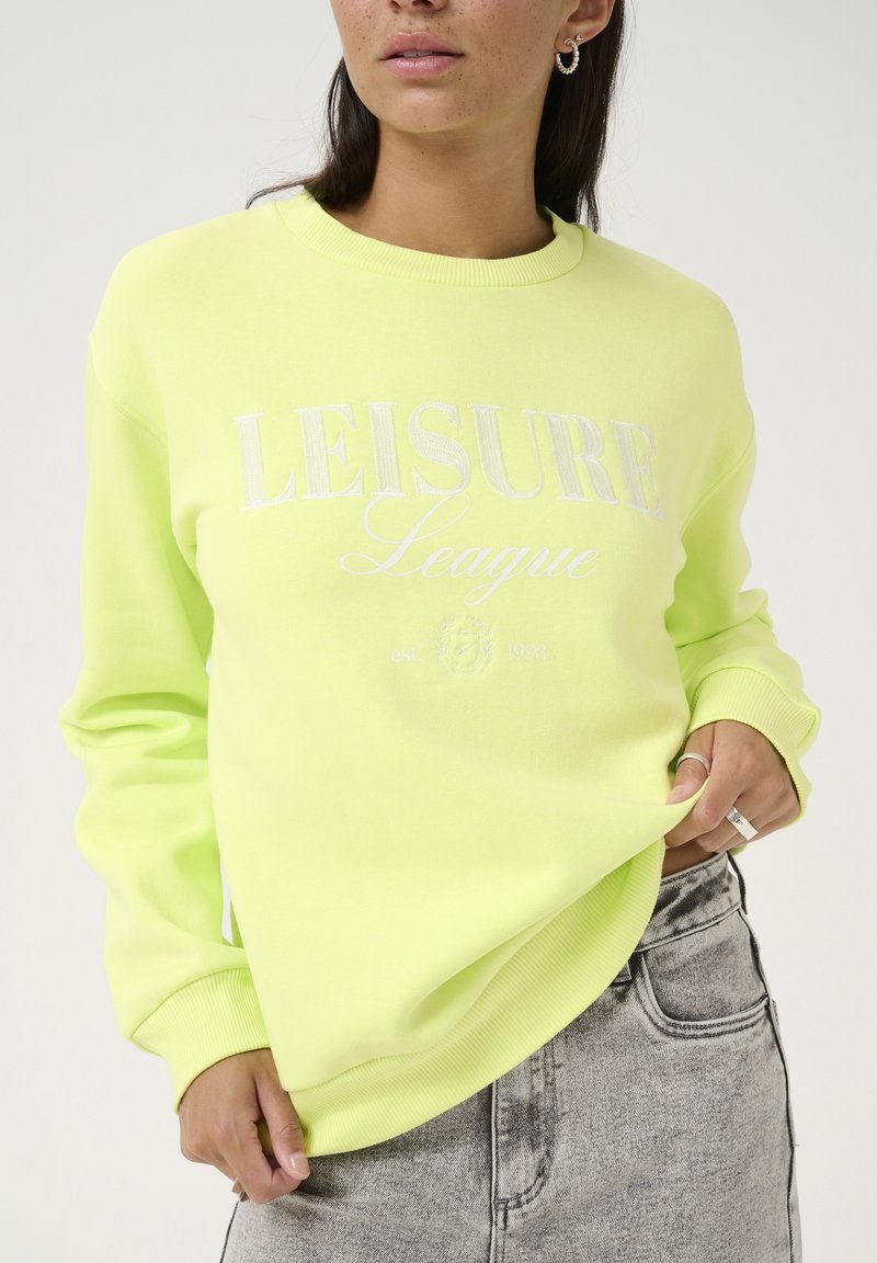 Femme portant un sweat-shirt jaune clair avec le texte "LEISURE League", associé à un jean gris délavé, visible des épaules jusqu'aux hanches.