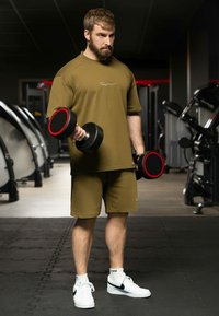 Tenue de sport vert olive composée d'un t-shirt ample et d'un short. Haltères noirs avec des accents rouges. Baskets blanches avec logo noir. Cadre de salle de sport.