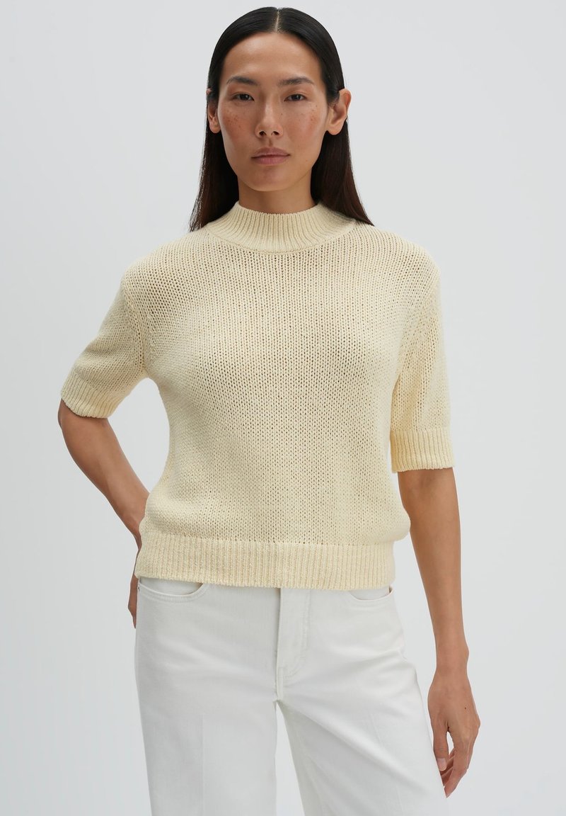 Frau mit langen dunklen Haaren, die einen kurzärmligen cremefarbenen Strickpullover und weiße Hose trägt und vor einem einfarbigen Hintergrund steht.