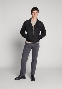 Black denim jacket with two chest pockets, worn over a light beige shirt, paired with gray jeans and black shoes. Simple design and texture.