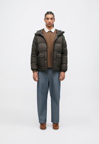 Polstret, mørkebrunt jakke med hette, frontlommer og glidelås. Kombinert med en brun genser, blå jeans og brune sko.