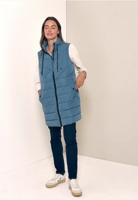 Gilet sans manches bleu clair, rembourré, avec fermeture éclair devant, capuche ajustable et motif matelassé horizontal. Associé à un jean foncé et des baskets.