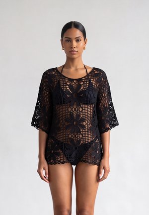 Donna con un copricostume nero a uncinetto floreale sopra un bikini nero, in piedi con le braccia rilassate lungo i fianchi contro uno sfondo chiaro e semplice.
