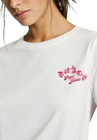 Camiseta de algodón blanca con un estampado gráfico rojo en el lado izquierdo del pecho que incluye el texto "Pepe Jeans" y un motivo de lazo. Cuello redondo estándar.