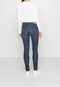 Tajta jeans i mörkblå denim med medelhög midja, med bakfickor och sydda detaljer, kombinerade med en vit långärmad topp och vita sneakers.