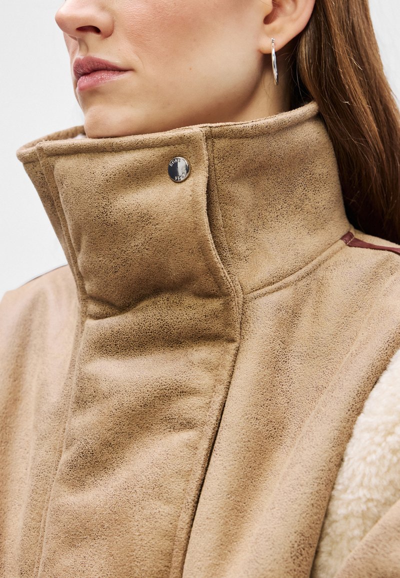 Tanssuedejacke mit hohem Kragen, silbernen Druckknopfdetails und einer weichen Textur sowie einem kontrastierenden Innenstoff.