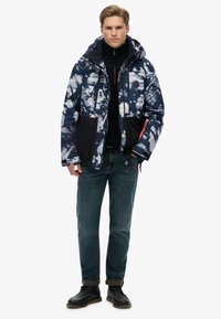 Polstret jakke i marineblå med abstrakte hvid og grå mønstre, sorte accenter, frontlommer og hætte, parret med blå jeans og sorte sko.