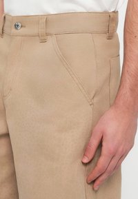 Μπεζ παντελόνι με διαγώνιο ραφές στις μπροστινές τσέπες, ασημένιο κουμπί κλεισίματος και ένα χέρι να ακουμπά δίπλα στον δεξιό μηρό πάνω σε λευκό πουκάμισο.