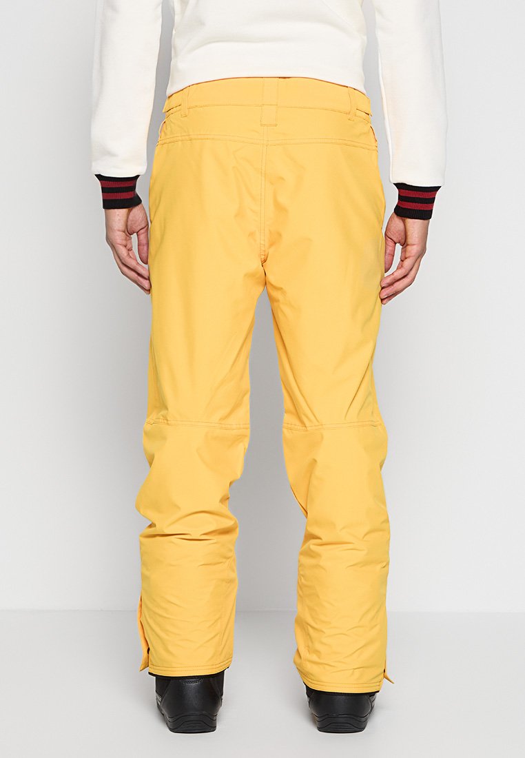 Gelbe Skihosen mit geradem Bein, strapazierfähigem Material und Reißverschlüssen an den Knöcheln. Der Bund hat Gürtelschlaufen und eine glatte Oberfläche.