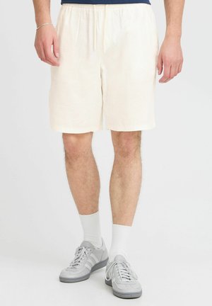 Cream-colored linen shorts with an elastic waistband, falling to mid-thigh, paired with gray sneakers and white socks.