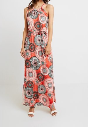 Femme portant une robe maxi corail sans manches avec des motifs floraux circulaires et une ceinture à la taille, associée à des sandales blanches à bouts ouverts.