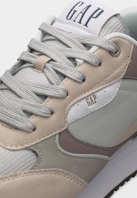 Κοντινό πλάνο μπεζ και γκρι αθλητικού παπουτσιού με γκρι κορδόνια, υφές σουέντ και διχτυού, και λογότυπο GAP στη γλώσσα και στην πλευρική επιφάνεια.