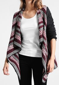 Stickad sjalcardigan i burgunderröd, svart och vit. Har en öppen front med ett mönstrat design, draperad asymmetrisk nederkant och långa ärmar.