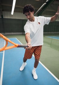 Maglietta bianca a maniche corte con logo, pantaloni cortes arancioni a quadri, in mano una racchetta da tennis arancione su un campo blu con sfondo verde.