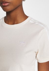 Detailní záběr na osobu v krémovém tričku Adidas s logem a třemi pruhy na rameni, doplněném o stříbrný náušník.