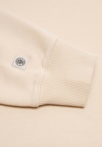Beige sweat top featuring ribbed cuffs and hem, made of soft fabric with a small woven label near the cuff detailing the brand.