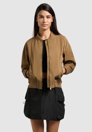 Kobieta o długich ciemnych włosach, ubrana w brązową bomberkę na zamek na czarnym krótkim topie i czarnej spódnicy typu cargo, stojąca z rękami w kieszeniach.