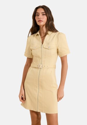 Kobieta w beżowej, krótkiej sukience z paskiem, zamkiem błyskawicznym z przodu i kieszeniami na piersi, stojąca na jednolitym jasnym tle.