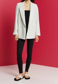 Blazer gris claro de corte cruzado con solapas de muesca, combinado con pantalones negros ajustados y sandalias de punta abierta con una banda cruzada blanca.