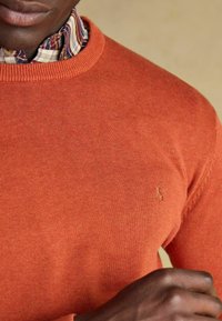Oranžový pletený sveter s okrúhlym výstrihom, s jemným logom na hrudi. Pod ním je čiastočne vidieť károvanú košeľu pri golieri.