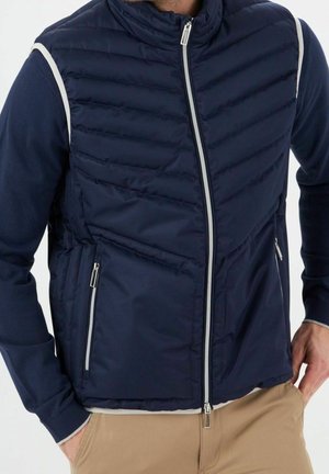 Homme portant une veste matelassée zippée bleu marine sur une chemise à manches longues avec un pantalon beige, les mains dans les poches, sur fond blanc.