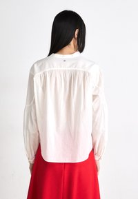 White cotton long-sleeve shirt with a relaxed fit, gathered back, and buttoned cuffs. Features a small logo at the nape of the neck.