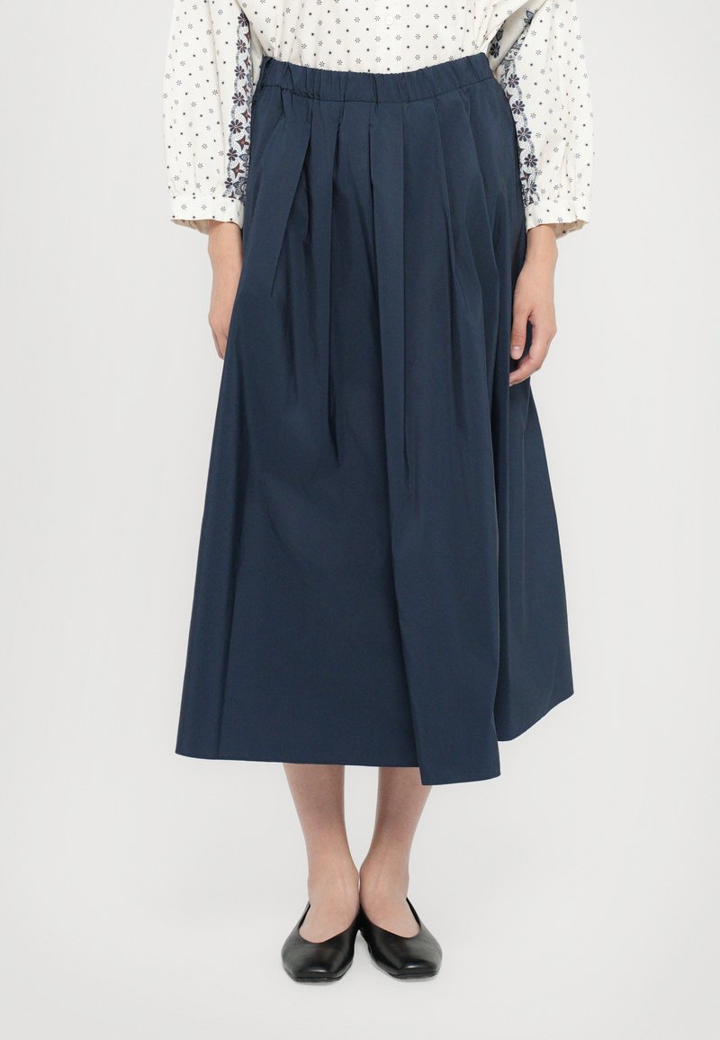Marineblauer Faltenrock aus leichtem Stoff; hat einen elastischen Bund, ist knöchellang und hat eine glatte Textur. Getragen mit schwarzen Ballerinas.