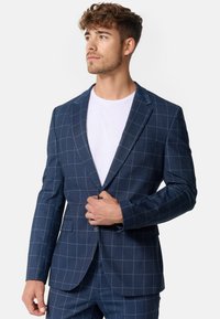 Marineblå ternet blazer med en enkelt knap, der har tekstureret stof, en spids krave og to frontlommer, båret over en hvid skjorte.