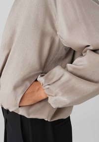 Licht beige satijnen top met lange mouwen, gathered manchetten en zijkleeddetails. De stof heeft een subtiele glans en een zachte textuur.