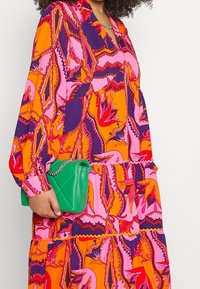 Kleurvolle jurk met lange mouwen en een levendig bloemenpatroon in oranje, roze en paars. Een kleine groene quilted handtas wordt in de hand gehouden.