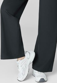 Osoba w czarnych, szerokich spodniach i białych, sportowych sneakersach, z nogami skrzyżowanymi na jednolitym, jasnym tle.