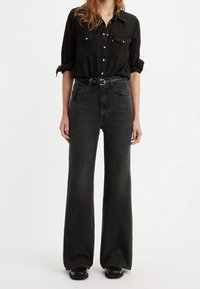 Femme portant une chemise noire boutonnée avec manches retroussées, un jean noir taille haute à jambes larges, une ceinture tressée noire et des chaussures noires.