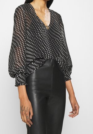Femme portant un chemisier noir transparent avec des motifs de traits blancs et un pantalon en cuir noir taille haute, accessoirisée d’un collier pendentif doré.