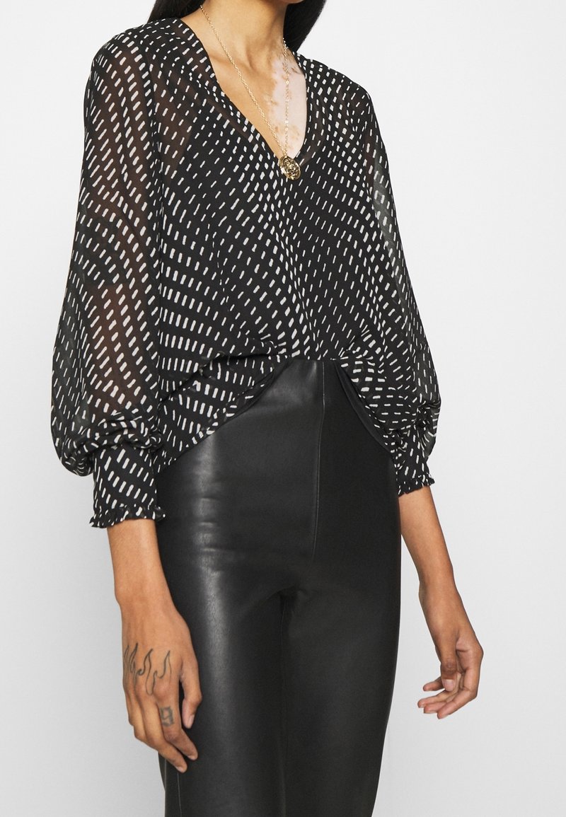 Femme portant un chemisier noir transparent avec des motifs de traits blancs et un pantalon en cuir noir taille haute, accessoirisée d’un collier pendentif doré.