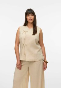 Beige mouwloos topje met een V-hals, voorzien van twee strikjes aan de voorkant. De stof heeft een gestructureerde, gekreukelde uitstraling. Gecombineerd met beige wijde broek.