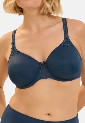 Femme portant un soutien-gorge bleu marine avec des bretelles brodées et un petit nœud au centre devant, montrée des épaules à la taille.